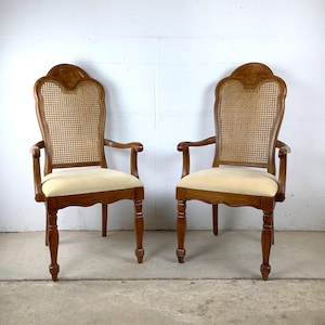 May include: A pair of wooden armchairs featuring high, arched backs with woven cane inserts, curved wooden frames, and cream-colored upholstered seats. The chairs have carved detailing on the top of the backrests and tapered legs.