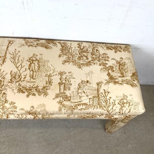May include: A rectangular bench with a cream-colored fabric featuring a brown toile pattern. The pattern depicts scenes of people in a garden setting. The bench has a padded top and a curved leg.