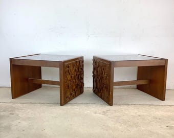 May include: Two matching wooden side tables. Each table has a dark top and a geometric carved design on the front. The tables are a medium brown color and have a simple, rectangular design.