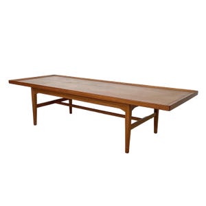 May include: A rectangular wooden coffee table with a light brown tabletop and a darker brown frame. The table has four angled legs and a lower support beam. The table measures approximately 60 inches long.