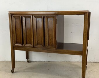 May include: A mid-century modern wooden bar cart with three cabinet doors and a lower shelf. The cart has a dark brown finish and is on wheels.