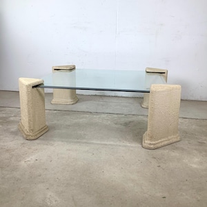 May include: A rectangular coffee table with a clear glass top and four beige, textured supports. The supports are rectangular with a flared base and a flat top, connected by a black metal bar. The table is a modern design.