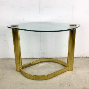 May include: A gold metal side table with a glass top. The table has a unique oval shape and a simple design.