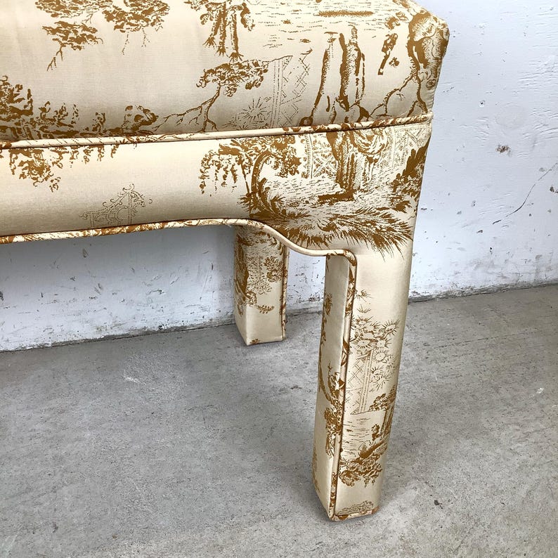 May include: A cream-colored bench with a toile pattern in shades of brown. The bench has a curved design and is upholstered with fabric featuring a detailed landscape scene. The legs are also covered in the same patterned fabric.