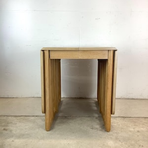 May include: A light-colored wooden side table with a rectangular top and vertical, grooved legs. The table has a simple, minimalist design, and the wood grain is visible. The table is approximately 30 inches tall.