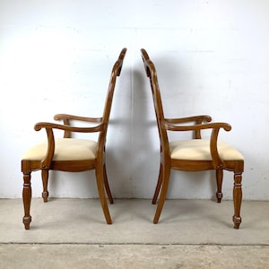 May include: Two wooden armchairs with high, curved backrests and light cream-colored upholstered seats. The chairs feature turned front legs, curved armrests, and a polished medium-brown wood finish, presented in a side-profile view.