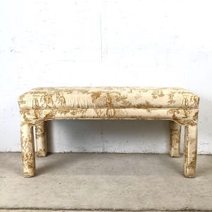 May include: A rectangular bench with a cream-colored fabric featuring a gold toile pattern. The bench has four short, square legs and a padded top. The fabric design includes figures, trees, and architectural elements.