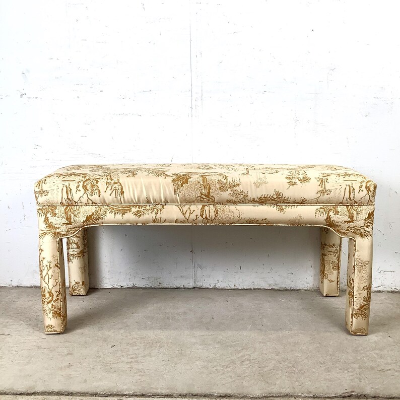 May include: A rectangular bench with a cream-colored fabric featuring a gold toile pattern. The bench has four short, square legs and a padded top. The fabric design includes figures, trees, and architectural elements.