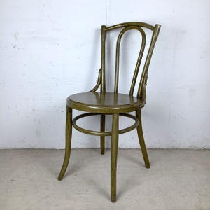 May include: A vintage olive green wooden chair with a rounded seat and curved back. The chair has four legs and a circular support near the base. The wood has a distressed finish, giving it a rustic appearance.
