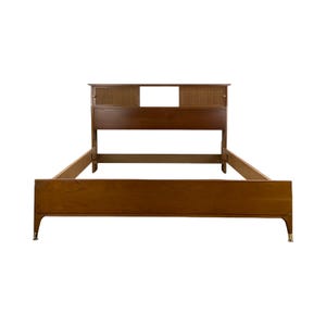 May include: A mid-century modern bed frame with a headboard featuring a woven cane insert and a rectangular cutout. The bed frame is made of wood with a warm brown finish and brass-colored feet. The bed frame is designed for a standard mattress.