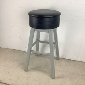 May include: A gray wooden bar stool with a black leather seat. The stool has a simple design with four legs and a single rung.