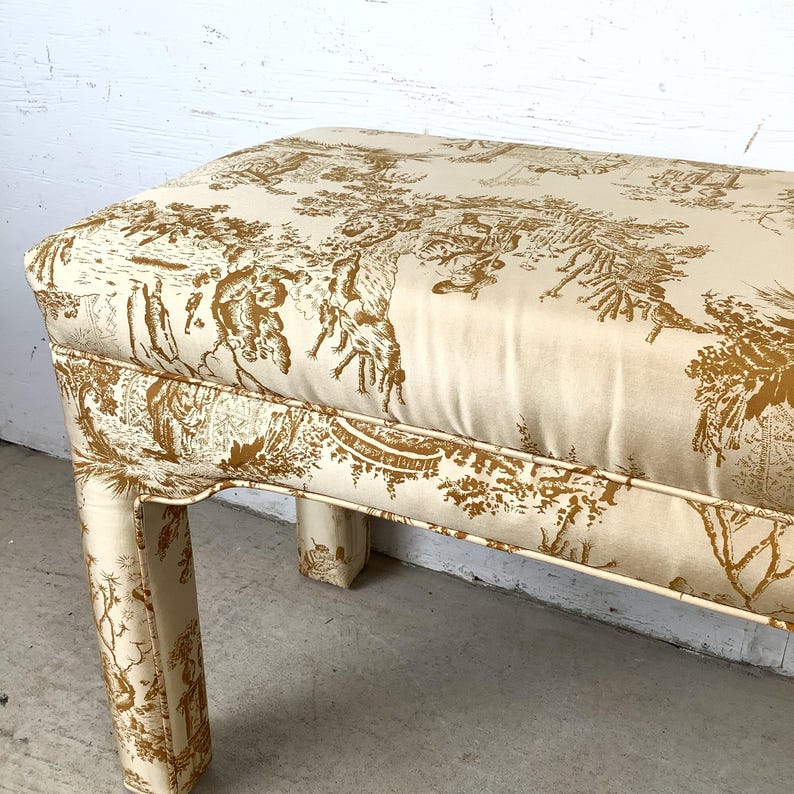 May include: Upholstered bench with a cream-colored background and a brown toile pattern depicting a pastoral scene. The bench has a rectangular seat and four curved legs. The fabric is tightly stretched over the frame.