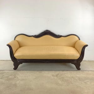 May include: A vintage-style sofa with a dark wood frame and a light yellow leather upholstery. The sofa has a curved back and a decorative carved design on the front.