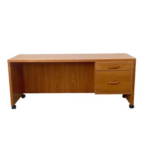 May include: A rectangular wooden desk with a light brown finish, featuring a flat top surface and a single column on the right side containing one small drawer above one larger drawer. The desk is supported by four small black caster wheels.