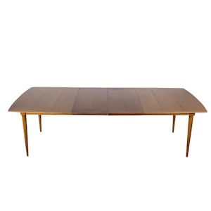 May include: A rectangular wooden dining table with a smooth, dark brown surface and a visible center seam indicating an extendable leaf. The table features four tapered, slender legs that angle outward from the corners, providing a stable base.