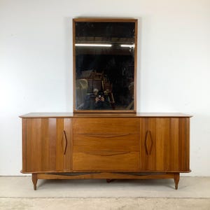 May include: A mid-century modern wooden dresser with a rectangular mirror. The dresser has two doors, three drawers, and a wooden frame. The mirror is rectangular with a wooden frame. The dresser is 60 inches wide.