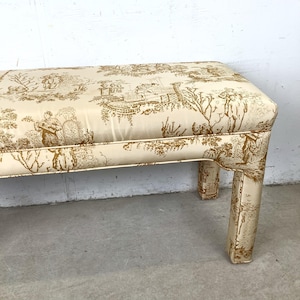 May include: A rectangular bench with a cream-colored fabric featuring a brown toile pattern. The bench has a padded top and four upholstered legs. The toile design depicts scenes of people, trees, and buildings.