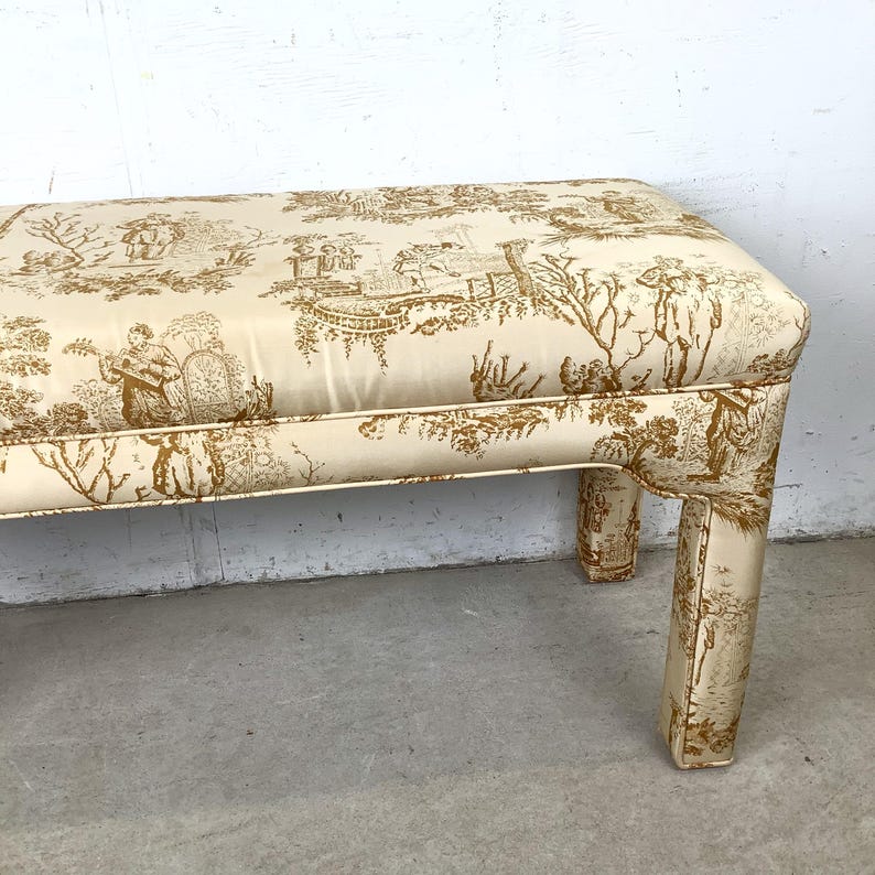 May include: A rectangular bench with a cream-colored fabric featuring a brown toile pattern. The bench has a padded top and four upholstered legs. The toile design depicts scenes of people, trees, and buildings.