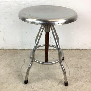May include: A vintage adjustable metal stool with a round, silver seat. The stool has a three-legged base and a threaded rod for height adjustment. The legs are silver with black rubber feet.