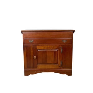 May include: A wooden cabinet with a warm, reddish-brown finish. It features a single drawer with ornate hardware, a cabinet door with a wooden knob, and decorative metal hinges. The cabinet has a classic, vintage design.