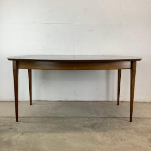 May include: A mid-century modern oval dining table with a wood top and tapered legs. The table is made of solid wood and has a rich brown finish.