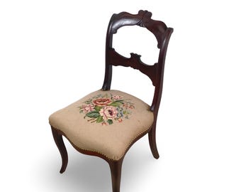 Puede incluir: Una silla de madera oscura con respaldo curvo y asiento tapizado en color beige que presenta un diseño central de bordado floral en tonos rosa, verde y azul, acentuado con pequeños clavos de latón en el borde del asiento.