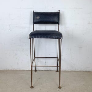 May include: A tall bar stool featuring a dark navy blue leather seat and backrest with visible stitching along the edges. The frame is constructed from thin, rusted brown metal rods that form four legs with circular feet and a square footrest.