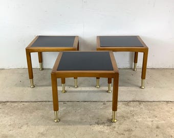 May include: Three square side tables with a dark, possibly black, top and wooden legs. The legs have brass-colored metal tips. The tables are made of wood and have a mid-century modern design.