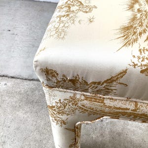 May include: Close-up of a cream-colored chair with a gold toile pattern. The fabric features detailed illustrations of landscapes and figures. The chair's design includes a curved seat and legs, showcasing a classic aesthetic.