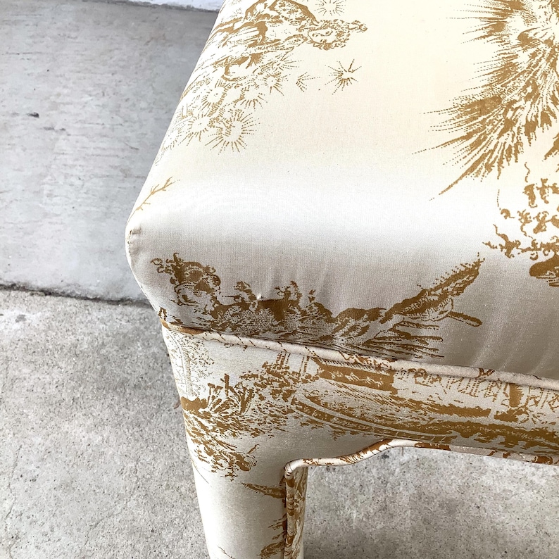 May include: Close-up of a cream-colored chair with a gold toile pattern. The fabric features detailed illustrations of landscapes and figures. The chair's design includes a curved seat and legs, showcasing a classic aesthetic.