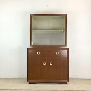 May include: A mid-century modern wooden cabinet with a glass-fronted display case on top. The cabinet has a warm brown finish, gold-toned square hardware, and a curved base. The display case has a shelf and a light-colored interior.