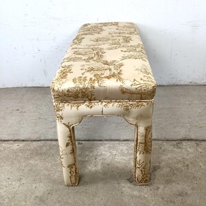 May include: A rectangular bench with a cream-colored base and a seat upholstered in a cream fabric with a gold toile pattern. The bench has four legs with a decorative cutout design.