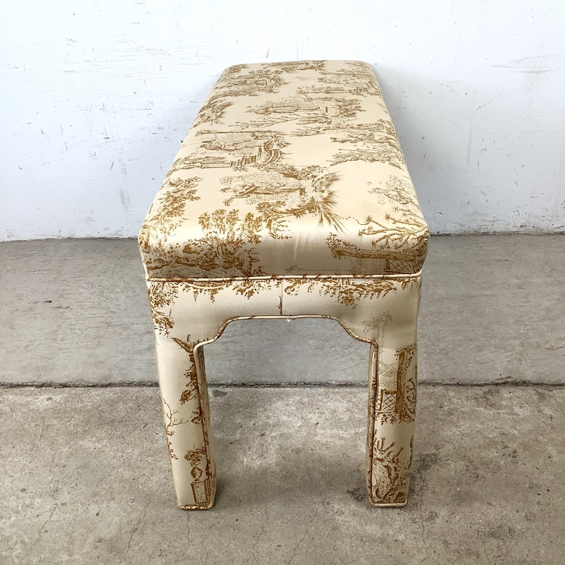 May include: A rectangular bench with a cream-colored base and a seat upholstered in a cream fabric with a gold toile pattern. The bench has four legs with a decorative cutout design.