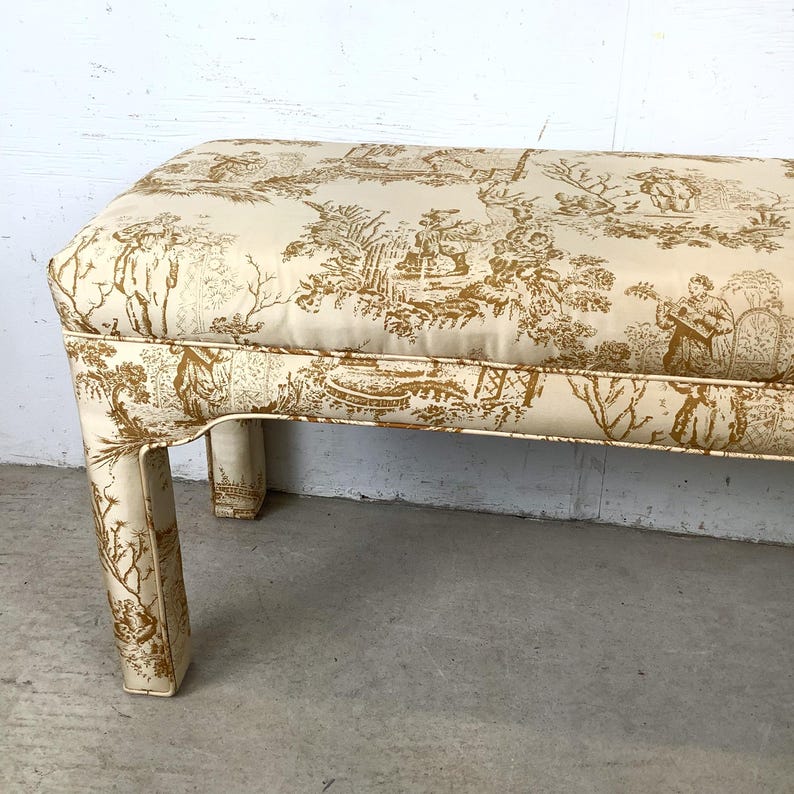May include: A rectangular bench with a cream-colored fabric featuring a gold toile pattern depicting pastoral scenes. The bench has four short, curved legs and a padded seat. The fabric is trimmed with a thin gold border.