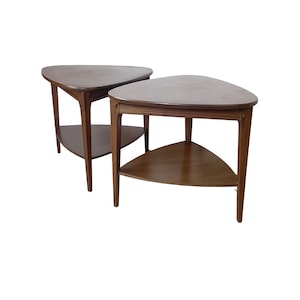 May include: Two matching wooden side tables. Each table has a triangular top and a lower shelf, supported by three legs. The tables are a medium brown color and have a mid-century modern design.