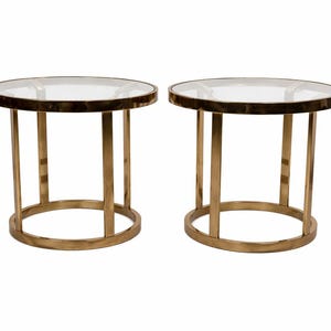 May include: Two round side tables with clear glass tops and brass-colored metal frames. Each table has a circular base, four vertical supports, and a circular rim around the glass top. The tables are identical.