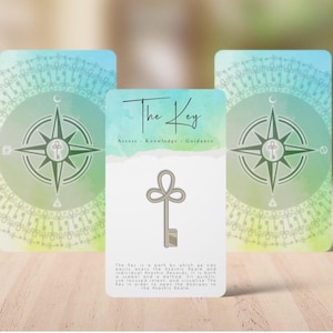 May include: Three tarot cards with a watercolor background. The center card features a key illustration and the words "The Key." The other two cards have a compass rose design. The cards are on a light wooden surface.