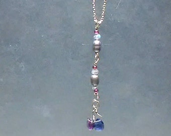 Pode incluir: Um delicado colar de prata com um pendente vertical. O pendente apresenta uma série de contas em prata, rosa e cores iridescentes, culminando em duas pedras preciosas azuis e roxas em forma de lágrima. A corrente é fina e o design geral é elegante.