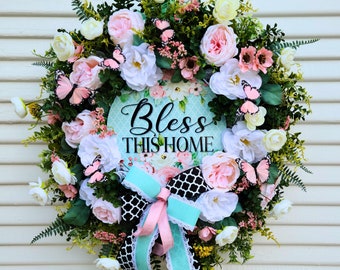 May include: A spring wreath featuring pink, white, and green flowers, butterflies, and greenery. The wreath has a sign that reads "Bless This Home" in black lettering on a turquoise background.