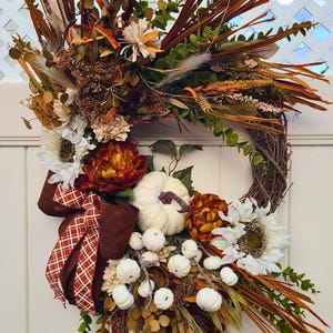 May include: A seasonal wreath featuring a grapevine base, decorated with white pumpkins, sunflowers, and dried flowers and foliage in orange, brown, and cream hues. A brown ribbon with a white plaid design adds detail.