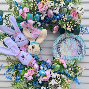May include: An Easter wreath featuring plush bunny faces, pastel eggs, and artificial flowers in pink, blue, and white. A round sign with bunny illustrations and "Happy Easter" is attached.