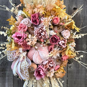 May include: A seasonal wreath with artificial flowers, pumpkins, and leaves. The wreath includes pink, burgundy, and peach-toned flowers, alongside small pumpkins in matching colours. It is decorated with dried grasses, leaves, and a ribbon with a leopard print.
