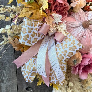 May include: A seasonal wreath with a large pink velvet pumpkin and gold stem, surrounded by faux flowers in pink, peach, and cream. The wreath is decorated with a bow made of white ribbon with gold leopard print and pink ribbon.