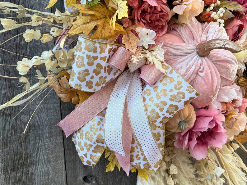 May include: A seasonal wreath with a large pink velvet pumpkin and gold stem, surrounded by faux flowers in pink, peach, and cream. The wreath is decorated with a bow made of white ribbon with gold leopard print and pink ribbon.
