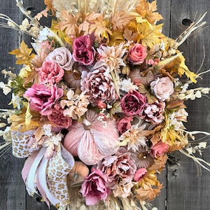 May include: A seasonal wreath with artificial flowers, pumpkins, and leaves in pink, peach, and gold hues. The wreath includes a decorative bow with a leopard print design. The arrangement is set against a rustic wooden backdrop.
