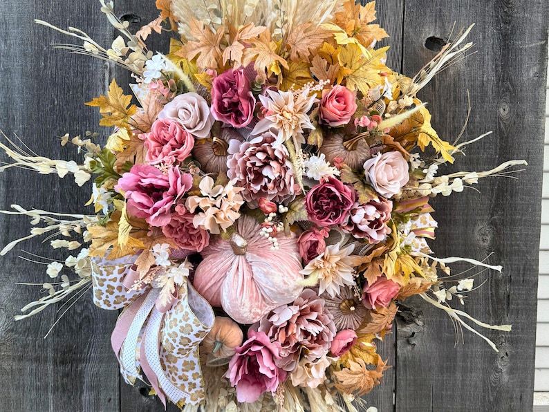 May include: A seasonal wreath with artificial flowers, pumpkins, and leaves in pink, peach, and gold hues. The wreath includes a decorative bow with a leopard print design. The arrangement is set against a rustic wooden backdrop.