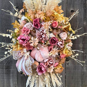 May include: A seasonal wreath featuring artificial flowers, pumpkins, and leaves in autumnal colours. The wreath includes pink, peach, and yellow blooms, with dried grasses and a decorative bow. It is displayed against a wooden backdrop.