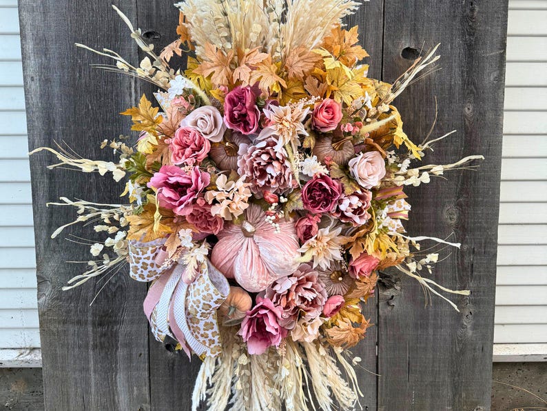 May include: A seasonal wreath featuring artificial flowers, pumpkins, and leaves in autumnal colours. The wreath includes pink, peach, and yellow blooms, with dried grasses and a decorative bow. It is displayed against a wooden backdrop.