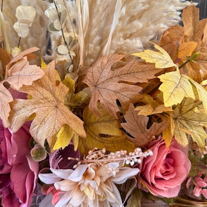 May include: Close-up of a seasonal floral arrangement. The display features large, colourful maple leaves in shades of yellow, orange, and brown. Pink and white roses and other flowers are also included, with dried grasses.