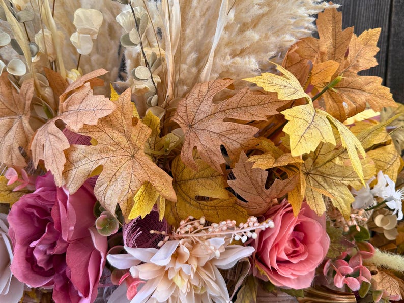 May include: Close-up of a seasonal floral arrangement. The display features large, colourful maple leaves in shades of yellow, orange, and brown. Pink and white roses and other flowers are also included, with dried grasses.